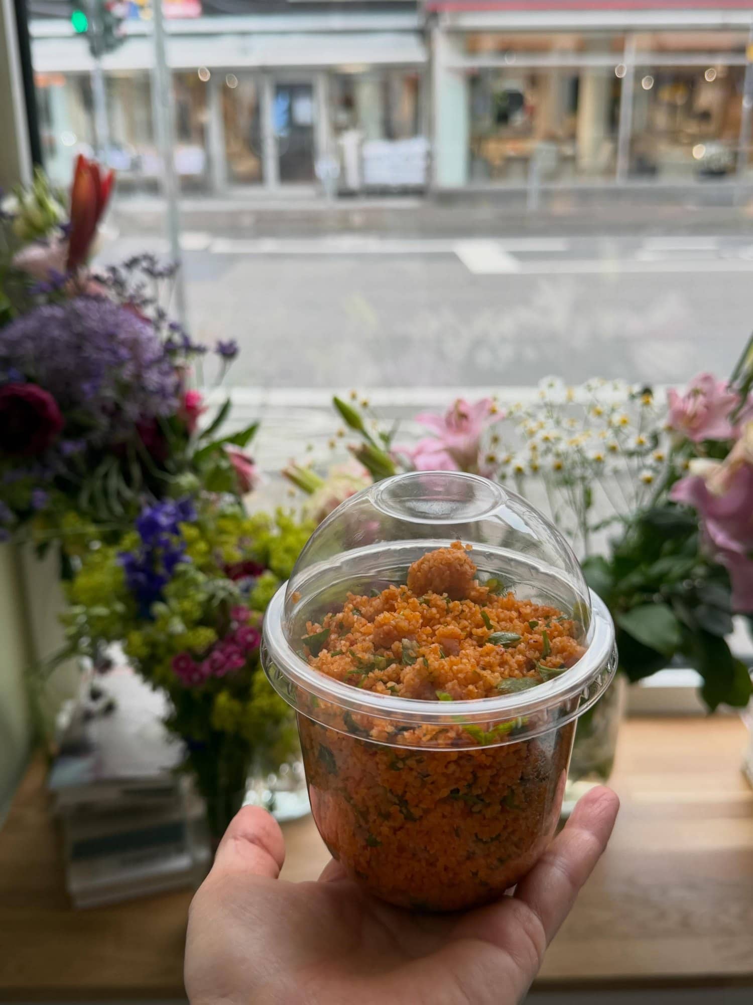Hausgemachter Kisir im Becher, vor dem Fenster zwischen bunten Blumen gehalten