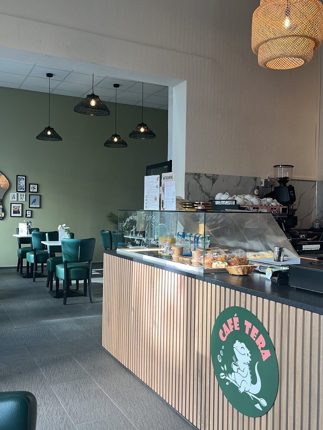 Innenraum von Café Tera: Theke mit Holzlamellen und Logo, Kuchenvitrine, dunkelgrüne Stühle und Rattanleuchten vor olivgrüner Wand